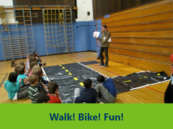 Image of a "Walk bike fun" activity in a school gym. An instructor is standing up and teaching to children sitting on the ground.